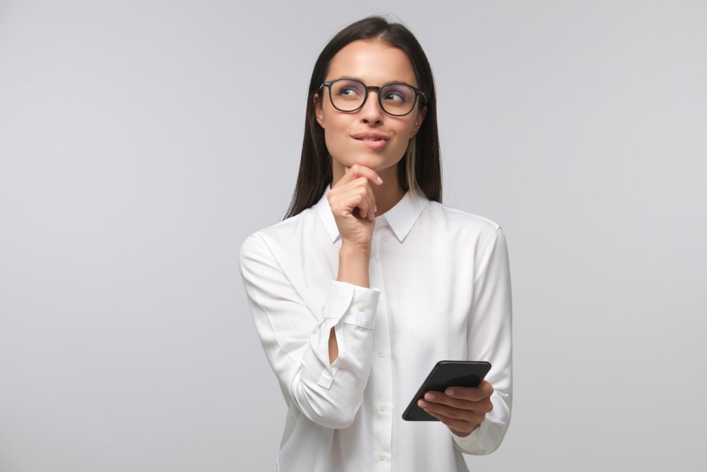 Young woman in white office shirt wearing eyeglasses and holding smartphone, making decisions and feeling doubt, looking aside, isolated on gray background