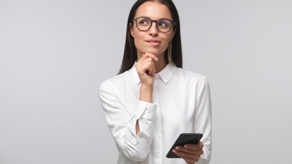 Young woman in white office shirt wearing eyeglasses and holding smartphone, making decisions and feeling doubt, looking aside, isolated on gray background
