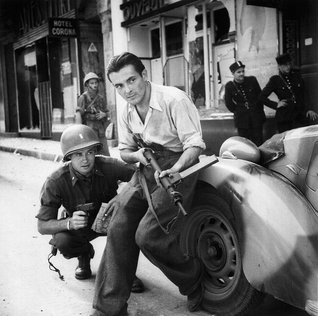 1030px-American_officer_and_French_partisan_crouch_behind_an_auto_during_a_street_fight_in_a_French_city._-_NARA_-_531322_-_restored_by_Buidhe