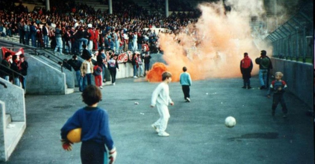 Деца играят с топки зад загражденията в един от секторите, а около тях се мятат факли, димки и бомбички по време на сблъсък между Тулуза и Ница в края на 80-те.