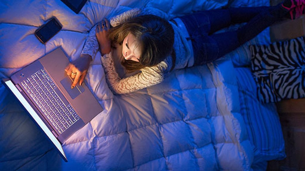 Teenage girl lying on a bed at night working on a laptop computer.