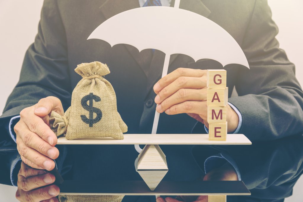 Businessman holds an umbrella to protect a US dollar money bag and cubes with a word GAME on a simple balance scale