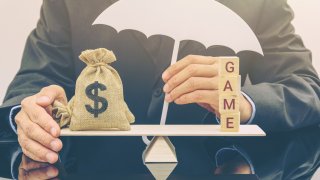 Businessman holds an umbrella to protect a US dollar money bag and cubes with a word GAME on a simple balance scale