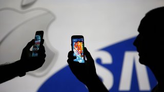 Men are silhouetted against a video screen with Apple and Samsung logos as he poses with Samsung S3 and Samsung S4 smartphones in this photo illustration taken in the central Bosnian town of Zenica