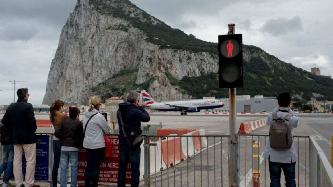 Хора чакат на светофара в Гибралтар да мине самолетът, за да отидат до отсрещната планина, която е известна забележителност.