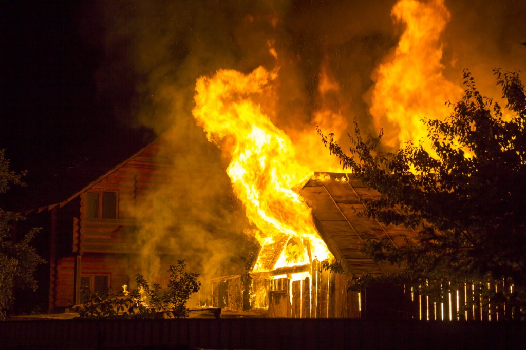 Burning wooden house at night. Bright orange flames and dense smoke from under the tiled roof on dark sky, trees silhouettes and residential neighbor cottage background. Disaster and danger concept.