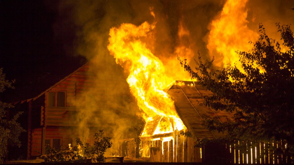 Burning wooden house at night. Bright orange flames and dense smoke from under the tiled roof on dark sky, trees silhouettes and residential neighbor cottage background. Disaster and danger concept.