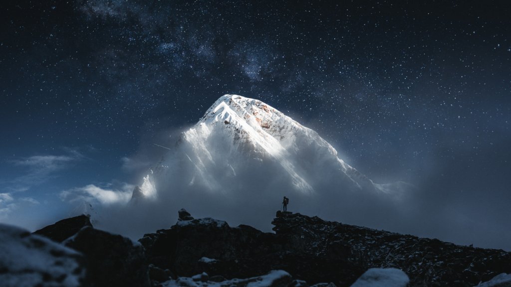 Brave solo mountain traveler stand in front of huge snowy mountain and starry sky. Professional tourist on mountains summit looking on greate snow cloudy rock