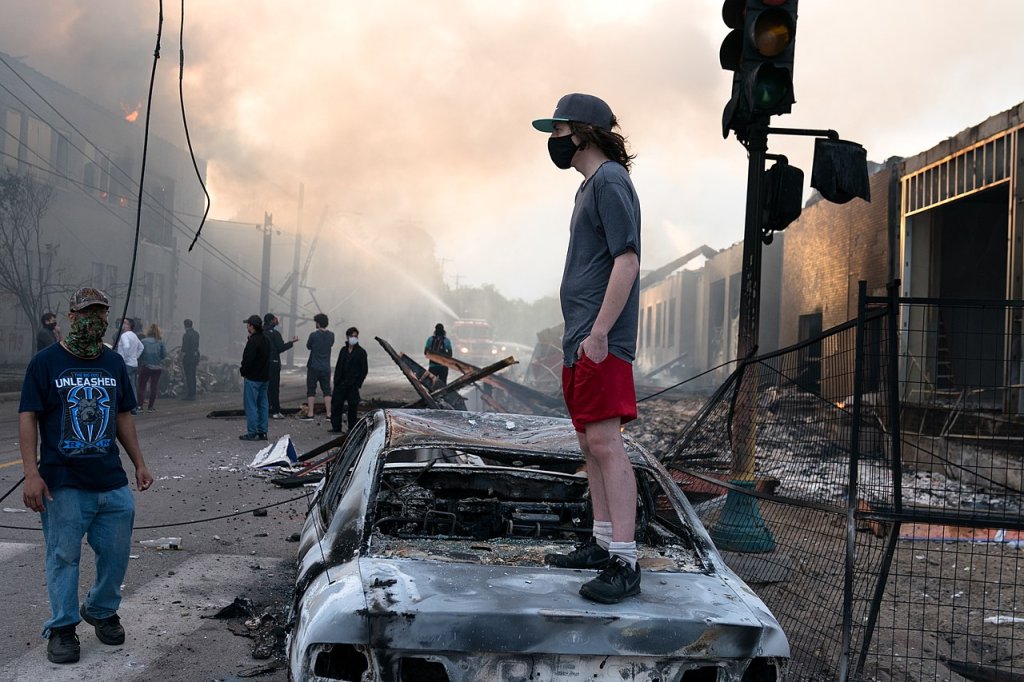 A_man_stands_on_a_burned_out_car_on_Thursday_morning_as_fires_burn_behind_him_in_the_Lake_St_area_of_Minneapolis,_Minnesota_(49945886467)