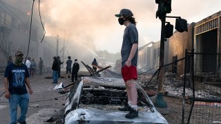 A_man_stands_on_a_burned_out_car_on_Thursday_morning_as_fires_burn_behind_him_in_the_Lake_St_area_of_Minneapolis,_Minnesota_(49945886467)
