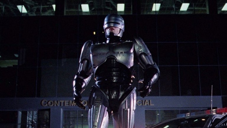 4. "Робокоп" (RoboCop, 1987)
Бъдещето не е особено приятно - консумеризмът е завладял обществото, престъпността вирее навсякъде, а властта е в ръцете на огромни корпорации. Алекс Мърфи (Питър Уелър) е полицай, който става жертва на жестоко (ама наистина ужасяващо) убийство. Мърфи възкръсва за нов живот, след като останките му са използвани за проекта "Робокоп" - създаването на комбинация между човек и машина, която по-ефективно да брани обществото. Но в този свят е трудно да определиш кои точно са "лошите".
Този киберпънк екшън е актуален и до днес, въпреки че е на повече от 30 години. Засяга теми като корупцията, авторитарната власт, влиянието на медиите, алчността и още. Има някаква ирония в частта за "алчността", защото филмът дава началото на цял франчайз от продължения, един римейк (задава се втори), анимационни и телевизионни сериали.
Верховен обаче няма общо с тях. Самият той първоначално е отвратен от сценария на филма, но с помощта на жена си вижда отвъд повърхността и възможността да се сатиризира американското общество (и сякаш неизбежното бъдеще, което вече може би е настояще).