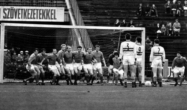 6 юни 1965 г. - Европейски приключения.
Флориан Алберт и Дежо Новак от Ференцварош мислят как да преодолеят стена от целия тим на Юнайтед в реванша  от полуфиналите на Купата на панаирните градове. "Червените дяволи" бият с 3:2 у дома, но губят с 0:1 в Унгария. Тогава правило за голове на чужд терен няма, та се налага 10 дни по-късно Юнайтед пак да пътува за Будапеща в трети мач. И отново губи. Европейската слава ще почака.
На снимката, от ляво на дясно: Джон Конъли, Шей Бренън, Ноби Стайлс, Денис Лоу, Боби Чарлтън, Били Фоулкс, Пат Креранд, Дейвид Херд, Джордж Бест, вратарят Пат Дън и Тони Дън.