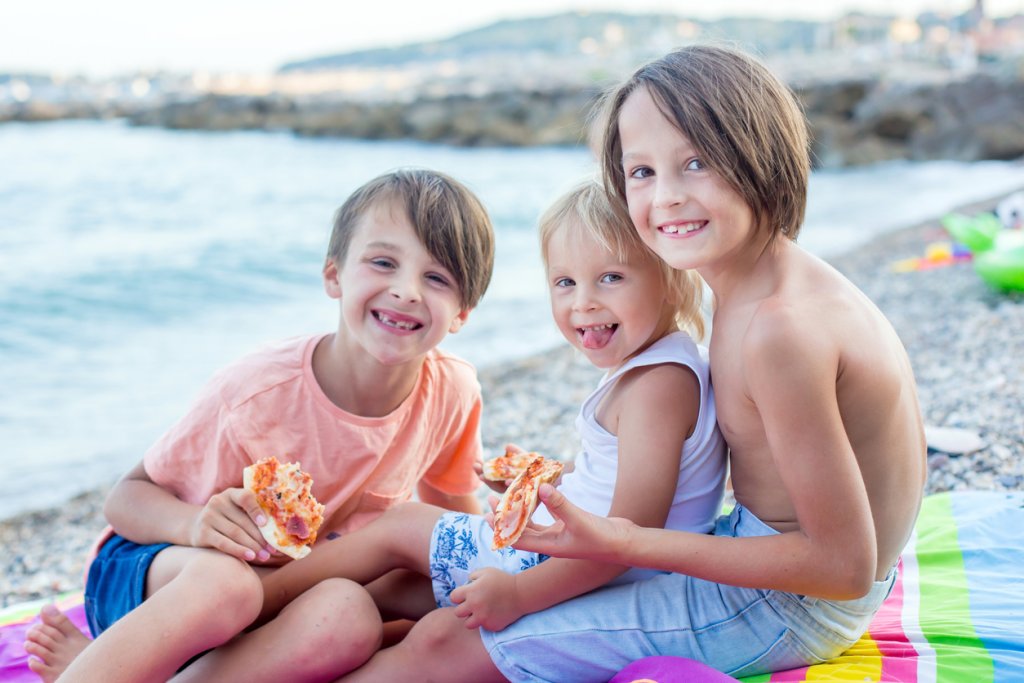 Sweet toddler child, eating pizza on the beach, having fun, smiling happily, kid enjoying dinner