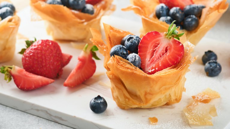 Сладки "кошнички" със сметана и плодовеТези „кошнички“ изглеждат много ефектно, ако се заиграем с декорацията им, но всъщност са елементарни за приготвяне. Готовите кори за баница се разстилат и се режат на шест правоъгълника. След това се разделят, мажат се с малко разтопено масло и се поставят във форма за мъфини така, че да се образуват „гнезда“.
„Кошничките“ се запичат до златисто на 170 градуса и се вадят да се охладят. През това време си приготвяме пълнежа от бита сметана, като в дълбока купа изсипваме голяма опаковка много студена течна сметана и половин чаша кристална захар. Бъркаме на най-високата степен на миксера и пълним „кошничките“. Украсяваме според наличните плодове и ядки.